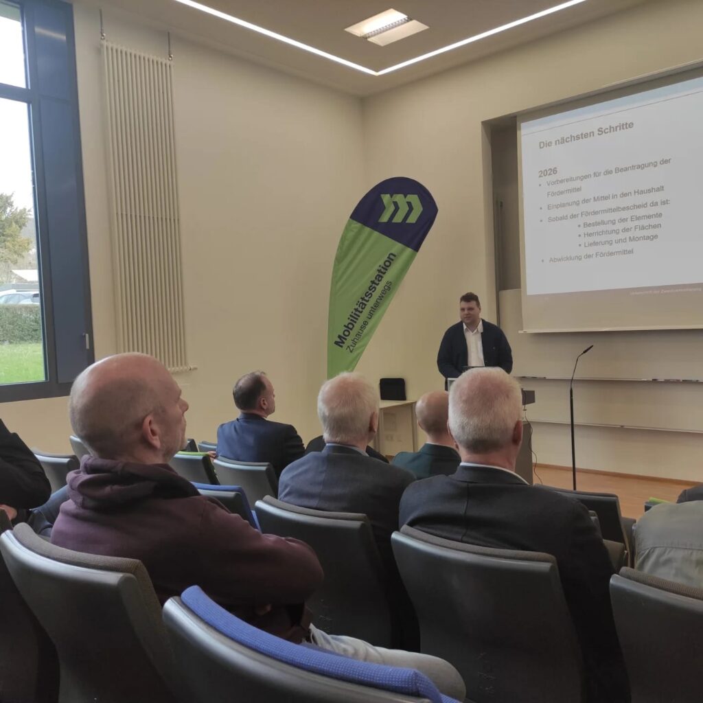 Ein Mann steht und präsentiert vor einem Publikum in einem Vortragssaal. Eine Präsentationsfolie wird an die Wand projiziert, und neben ihm ist ein grün blaues Banner mit der Aufschrift Mobilitätsstation zu sehen.